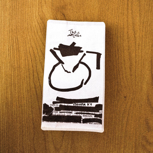 A rectangular white coffee bag with a black minimalist coffee maker illustration, placed on a brown wood-grain surface.